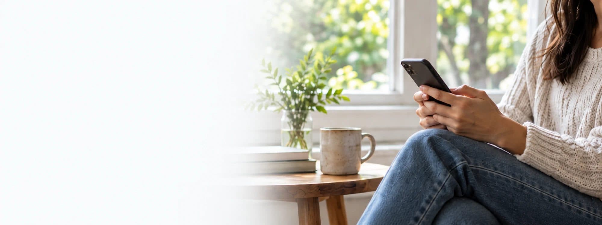 Someone sitting by a window with a phone, a mug and a small plant on the table beside them