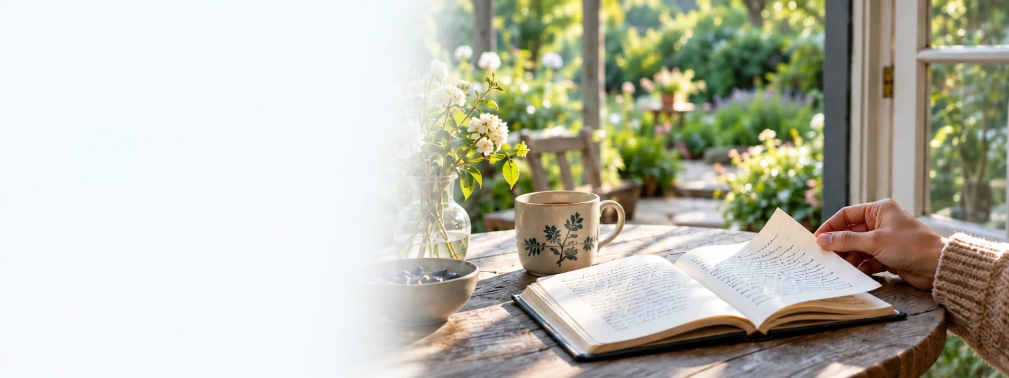 A notebook, a mug of tea and a jar of blossom on a garden table in morning light